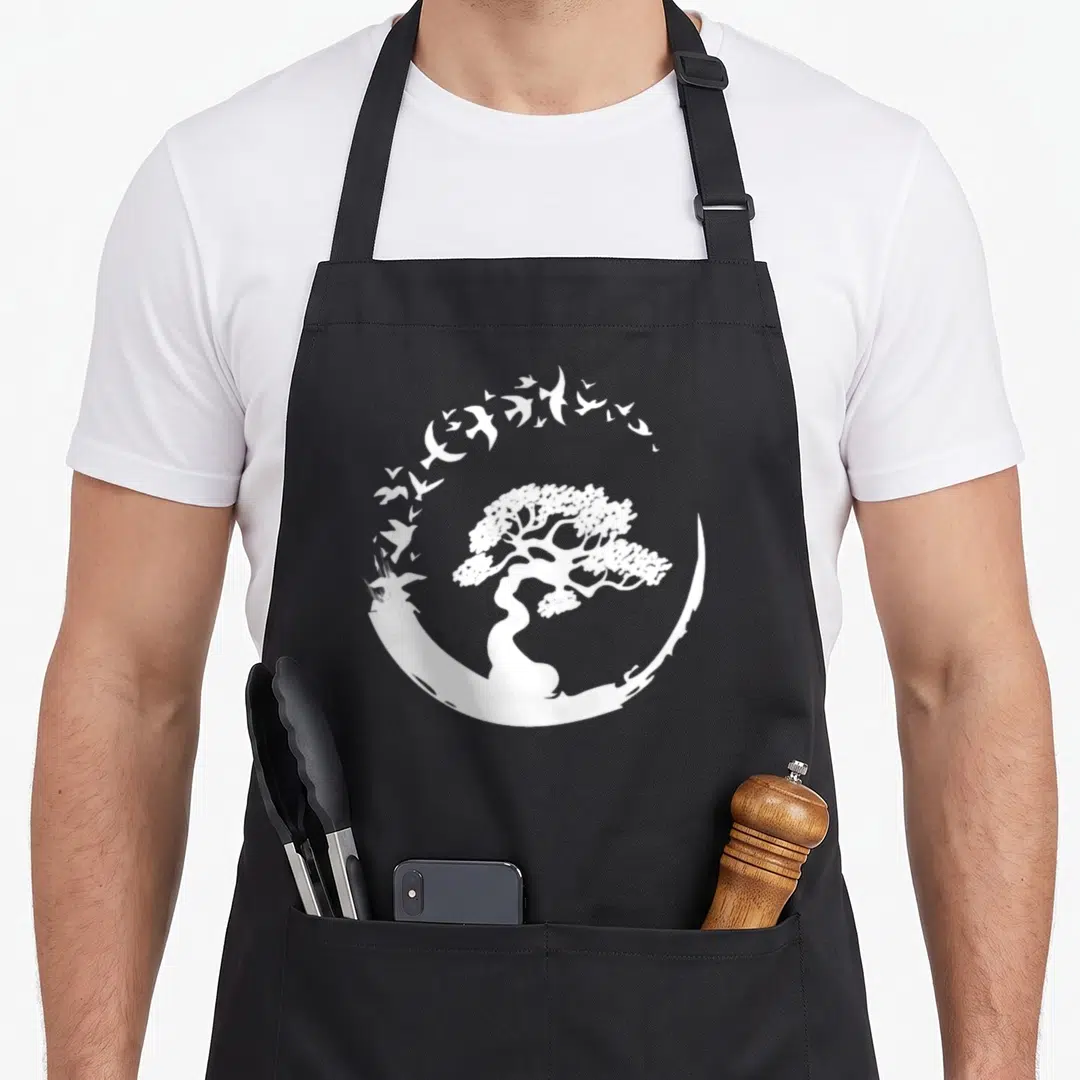 Japanese enso tree apron - White bonsai tree inside circular arc of flying birds, minimalist design on black apron — shown on a black cotton canvas apron against a white background