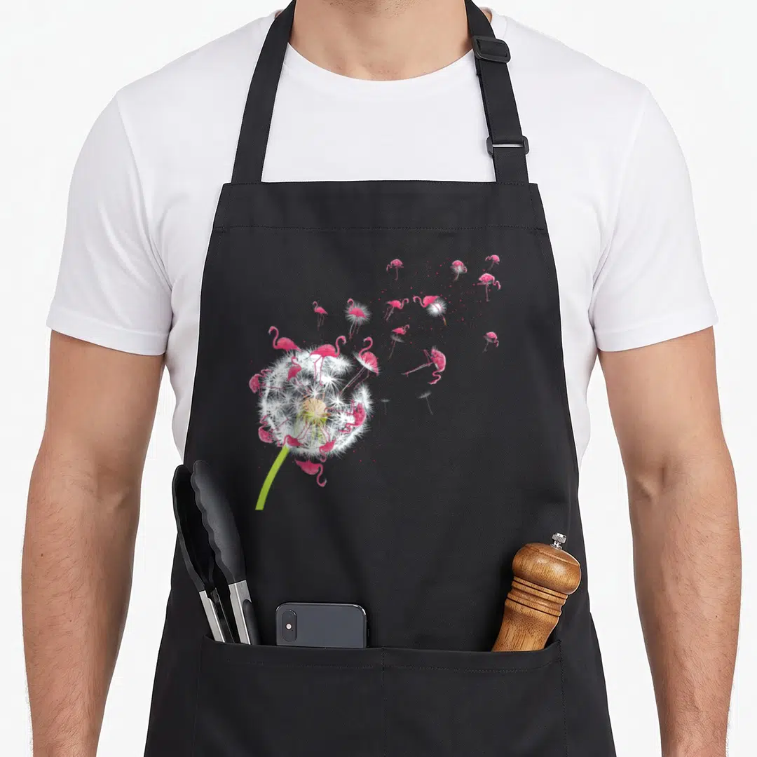 Tropical the floral apron - Pink flamingos blowing away from a white dandelion puff on a green stem — shown on a black cotton canvas apron against a white background