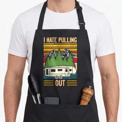 Pulling camping trailer apron - Retro camper trailer with I HATE PULLING OUT text and mountain sunset graphic — shown on a black cotton canvas apron against a white background