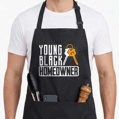Young housewarming apron - YOUNG BLACK HOMEOWNER text with two keys on a ring in white and orange — shown on a black cotton canvas apron against a white background