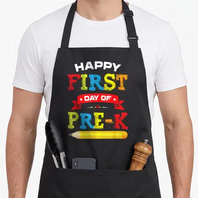 Student preschool apron - Stacked HAPPY FIRST DAY OF PRE-K text with red ribbon and yellow pencil illustration — shown on a black cotton canvas apron against a white background