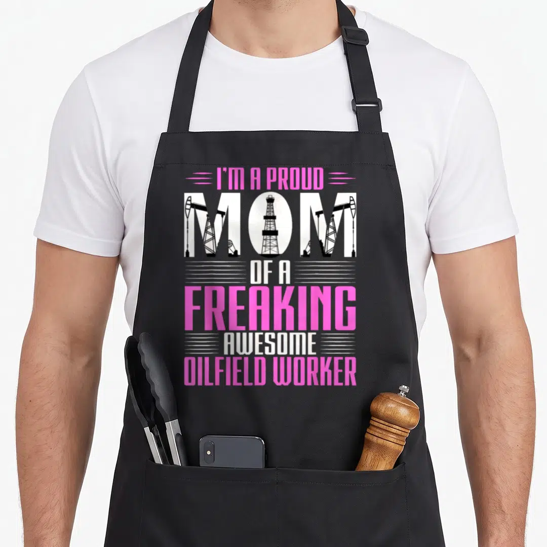 Rig oil mom apron - Pink and white typography with oil rig silhouettes inside the word mom and stacked text — shown on a black cotton canvas apron against a white background