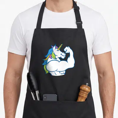 Muscular unicorn apron - Cartoon white unicorn flexing right arm with blue, yellow, pink, and orange mane — shown on a black cotton canvas apron against a white background