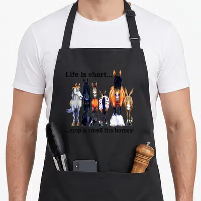 Stop horse apron - Cartoon horses and donkey in orange and brown with split text reading Life is short stop smell horses — shown on a black cotton canvas apron against a white background