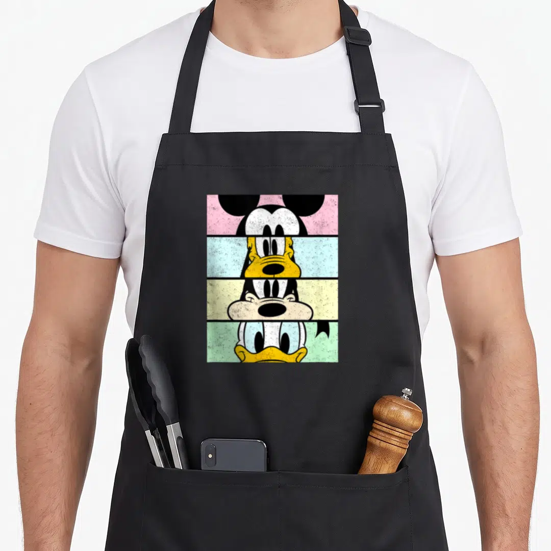 Comic friends cooking apron - Four stacked colored stripes forming a composite Disney face with retro texture and black — shown on a black cotton canvas apron against a white background