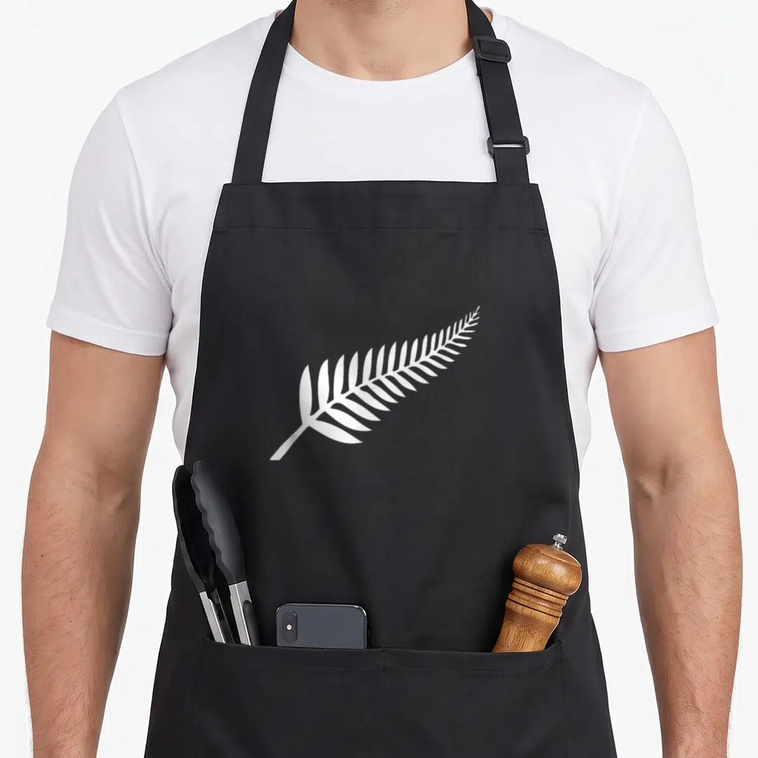 Pride new zealand apron - Single white fern leaf printed diagonally across the center of a dark apron panel — shown on a black cotton canvas apron against a white background