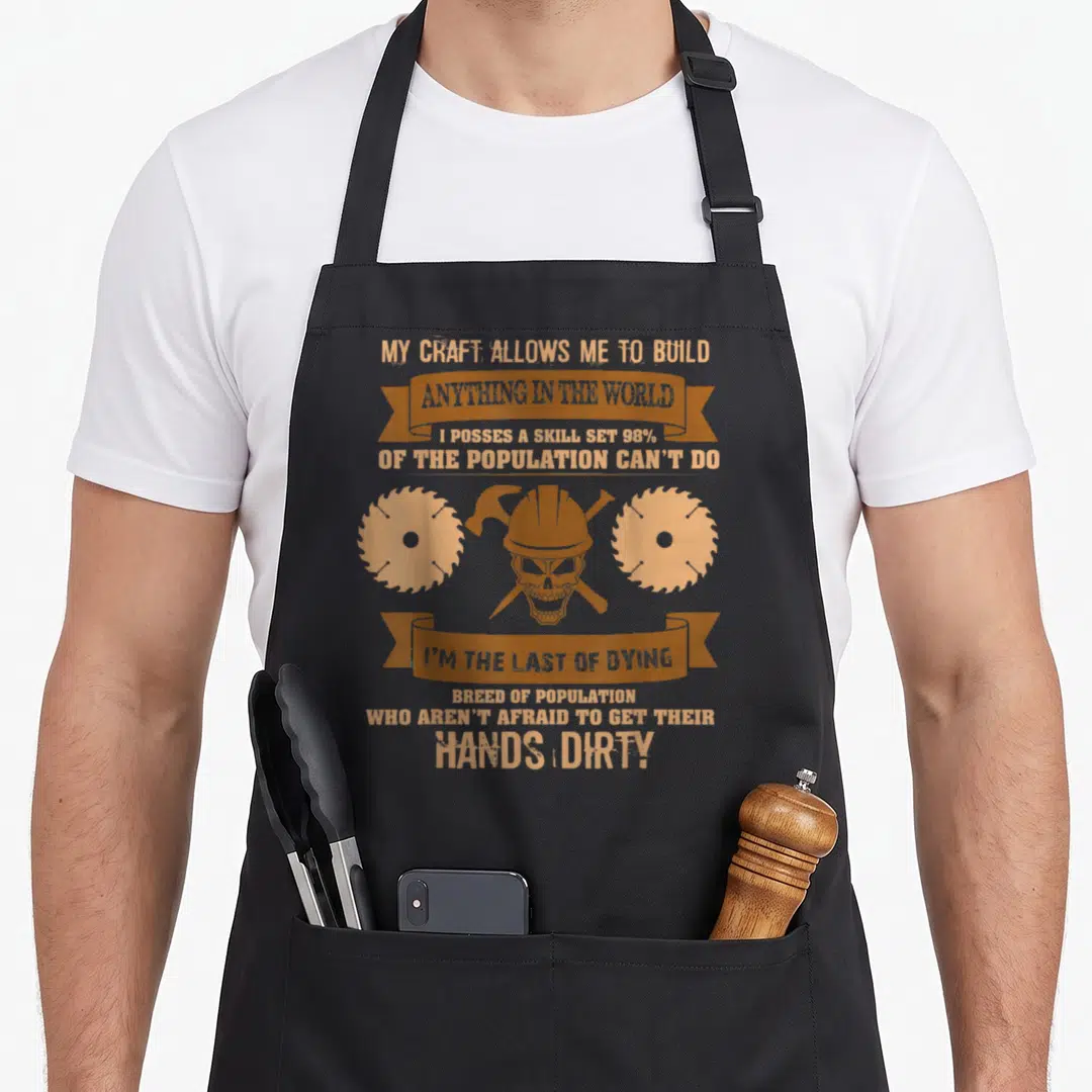 Vintage carpenter woodworking apron - Tan distressed skull in hard hat with hammer wrench and two saw blades flanking — shown on a black cotton canvas apron against a white background