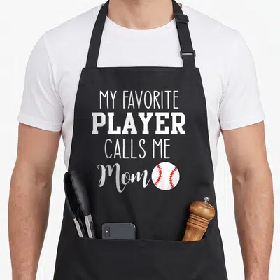 Cute player baseball apron - White text MY FAVORITE PLAYER CALLS ME Mom with a red-stitched baseball on the lower right — shown on a black cotton canvas apron against a white background