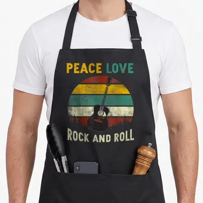 Rock peace apron - Black guitar over retro red yellow green striped sun with forest silhouette and PEACE LOVE ROCK — shown on a black cotton canvas apron against a white background