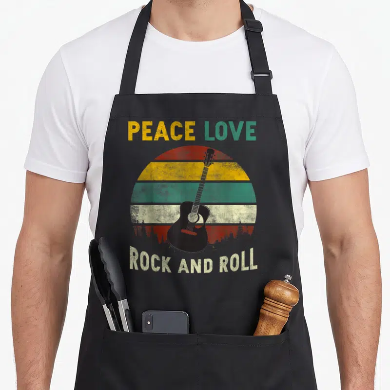 Rock peace apron - Black guitar over retro red yellow green striped sun with forest silhouette and PEACE LOVE ROCK — shown on a black cotton canvas apron against a white background