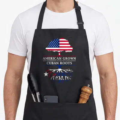 Grown cuban american apron - Tree with USA flag canopy and Cuba flag roots, split by text AMERICAN GROWN WITH CUBAN — shown on a black cotton canvas apron against a white background