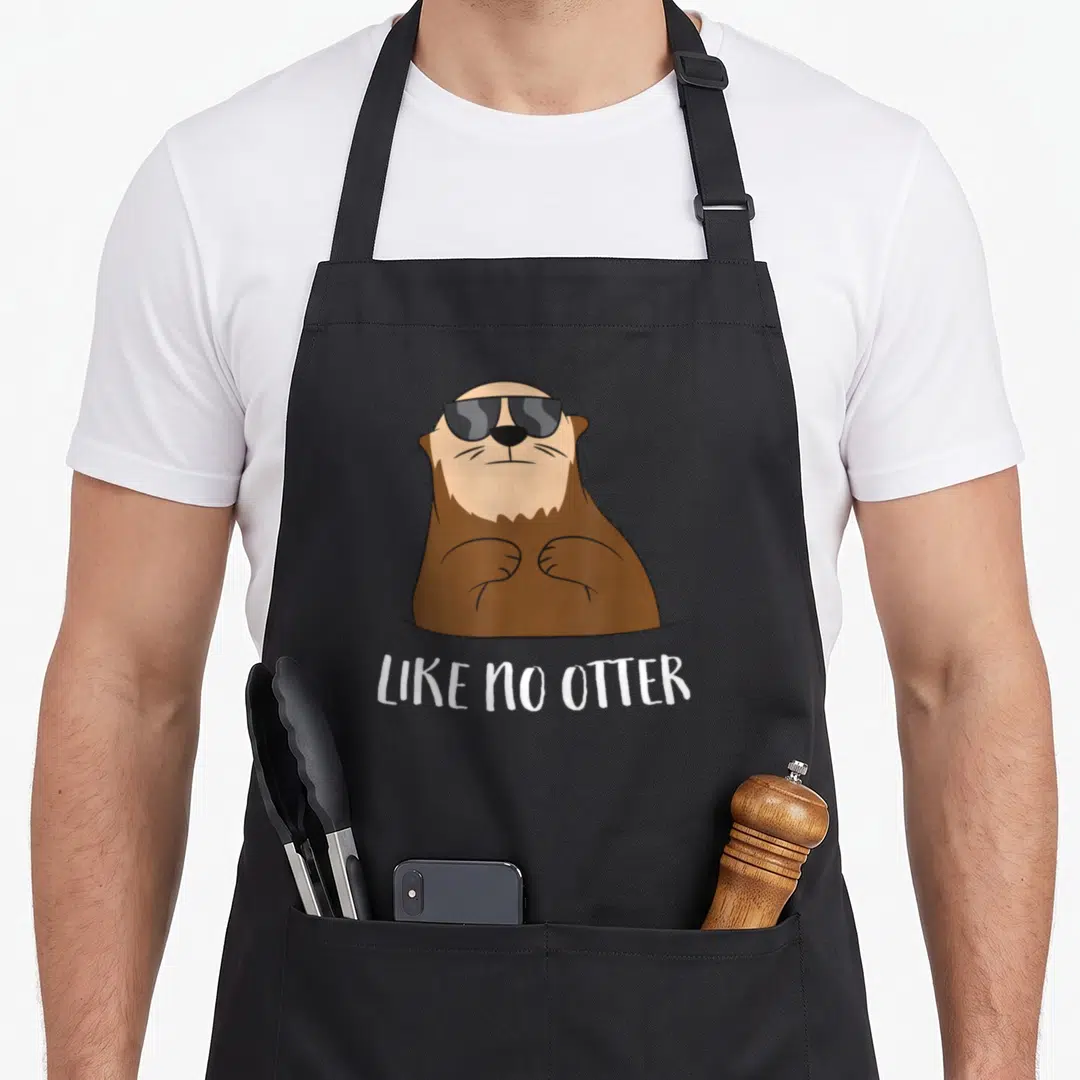 Retro sea otter apron - Cartoon otter in sunglasses above white text reading LIKE NO OTTER — shown on a black cotton canvas apron against a white background