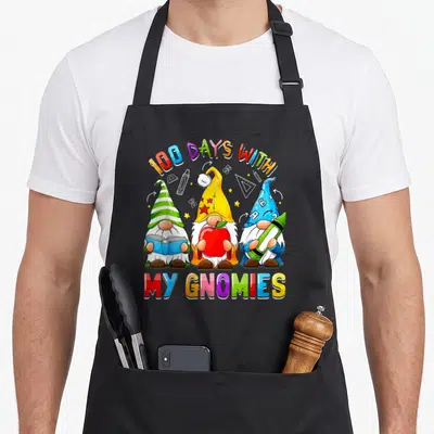 Gnome teacher apron - Three cartoon gnomes with school supplies under arching text 100 DAYS WITH MY GNOMIES — shown on a black cotton canvas apron against a white background