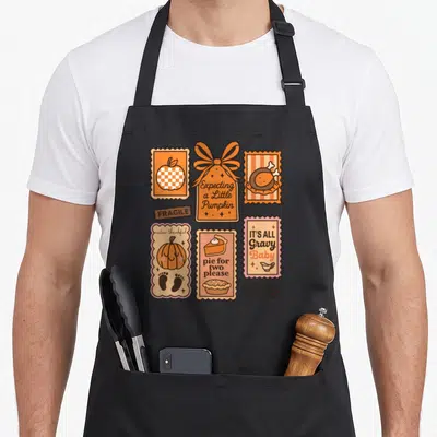 Maternity thanksgiving apron - Six vintage stamp graphics with pumpkin, pie, and pregnancy announcement text in orange — shown on a black cotton canvas apron against a white background