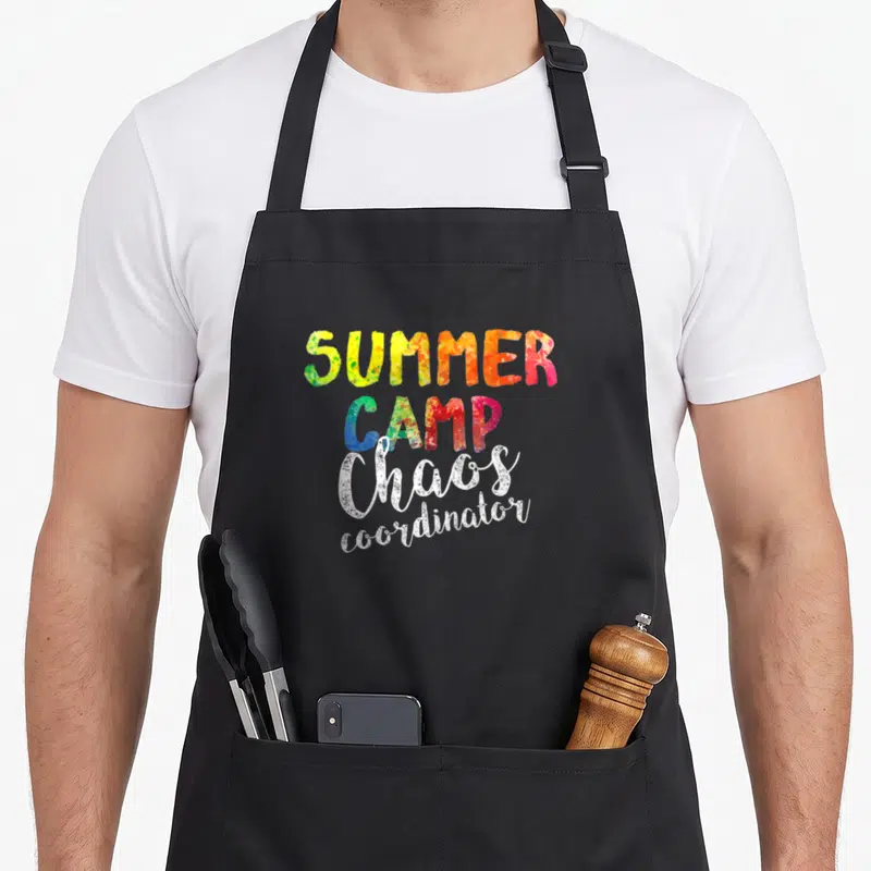Chao camp chef apron - Stacked text reading SUMMER CAMP Chaos coordinator in colorful speckled and white fonts — shown on a black cotton canvas apron against a white background