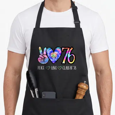 Senior tie-dye apron - Tie-dye peace sign heart and 76 with PEACE LOVE CLASS OF '76 text — shown on a black cotton canvas apron against a white background