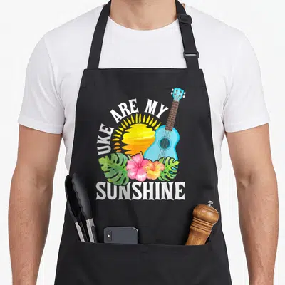 Sunshine ukulele apron - Watercolor blue ukulele, yellow sun, pink flowers, and Uke Are My Sunshine text — shown on a black cotton canvas apron against a white background