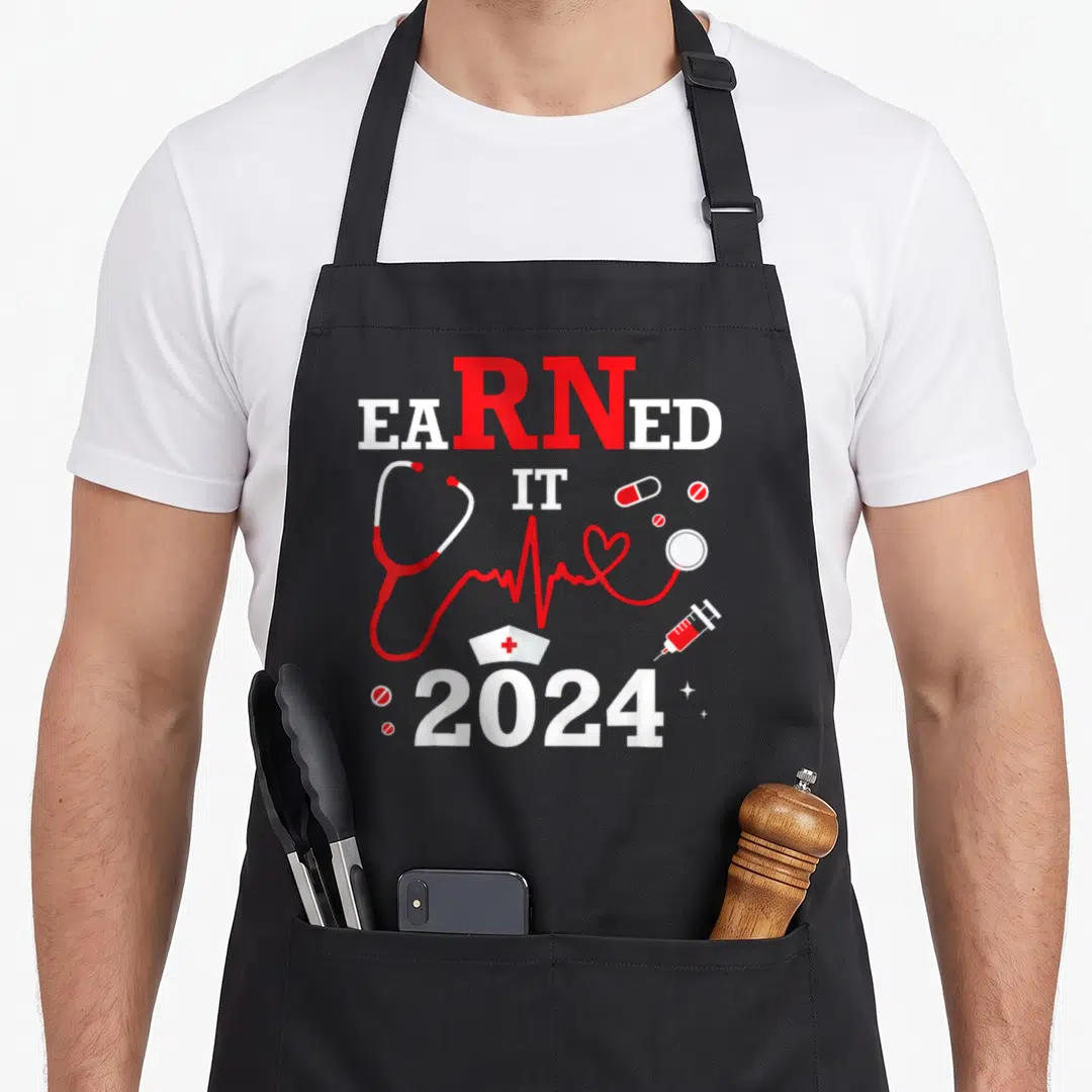 Earned nurse apron - EARNED IT 2024 text with red stethoscope heartbeat line nurse cap and medical tools — shown on a black cotton canvas apron against a white background