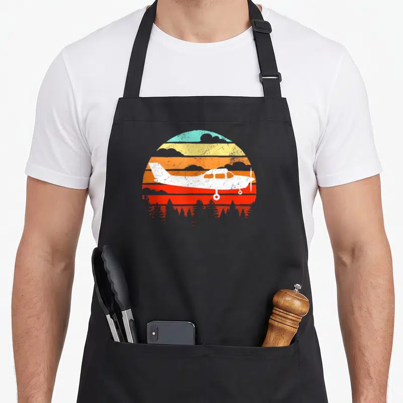 C172 aircraft apron - White airplane silhouette over retro striped sun with teal, orange, red, yellow bands and tree — shown on a black cotton canvas apron against a white background