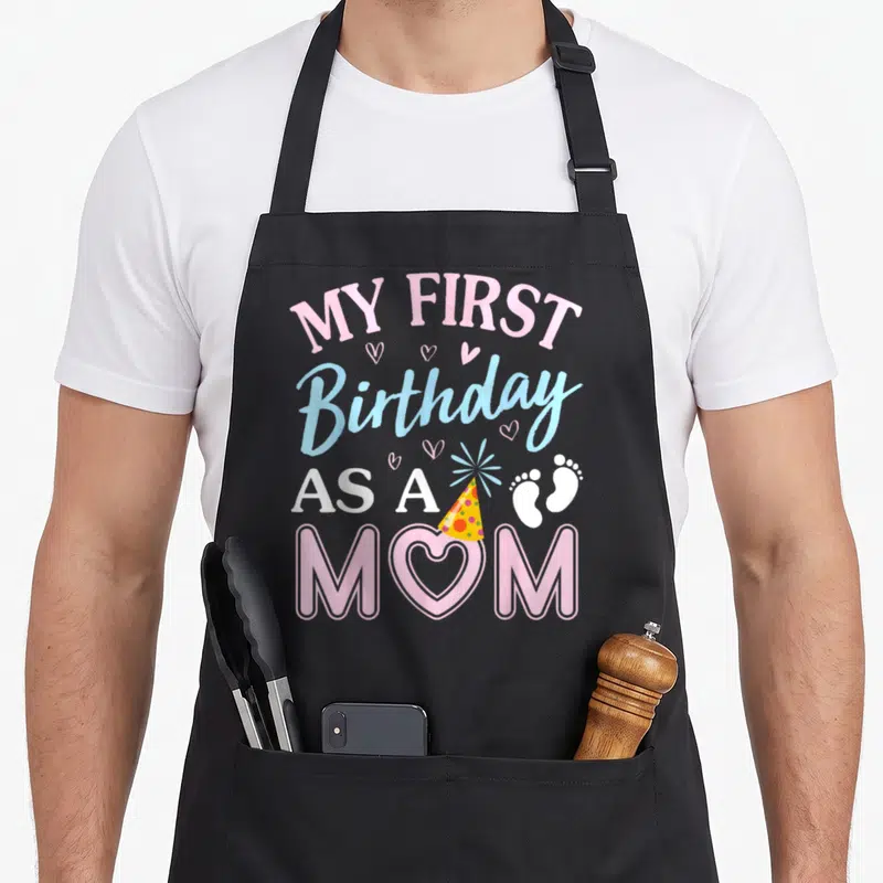 Birthday mama mother's day aprons - My First Birthday As A Mom text with heart, party hat, and footprints in pink — shown on a black cotton canvas apron against a white background