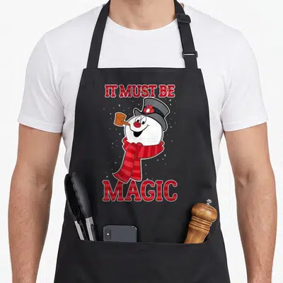 Frosty magic apron - Cartoon snowman with red scarf and IT MUST BE MAGIC text in red plaid font on grey background — shown on a black cotton canvas apron against a white background