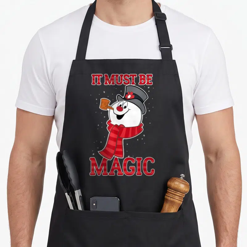 Frosty magic apron - Cartoon snowman with red scarf and IT MUST BE MAGIC text in red plaid font on grey background — shown on a black cotton canvas apron against a white background