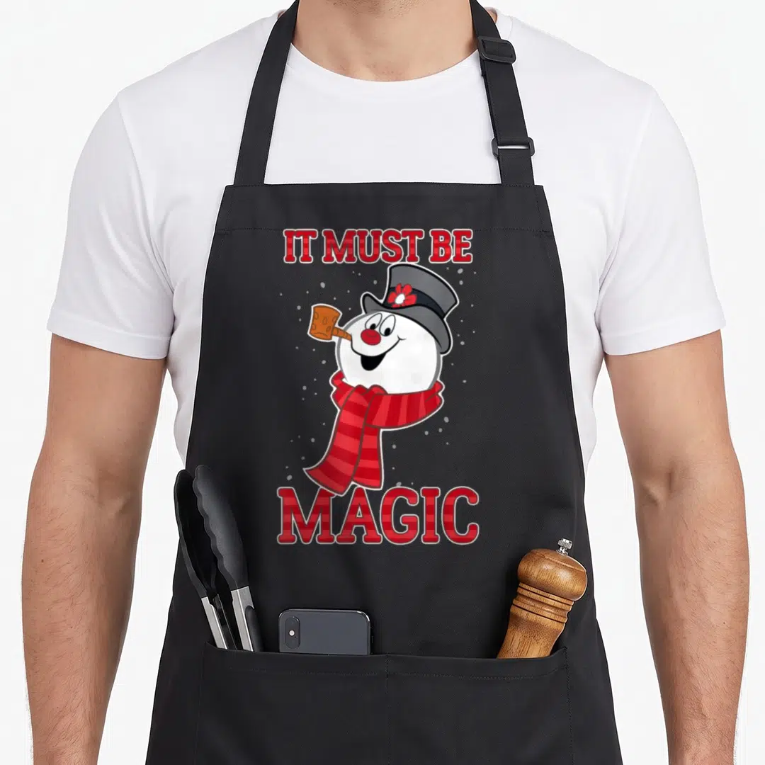 Frosty magic apron - Cartoon snowman with red scarf and IT MUST BE MAGIC text in red plaid font on grey background — shown on a black cotton canvas apron against a white background