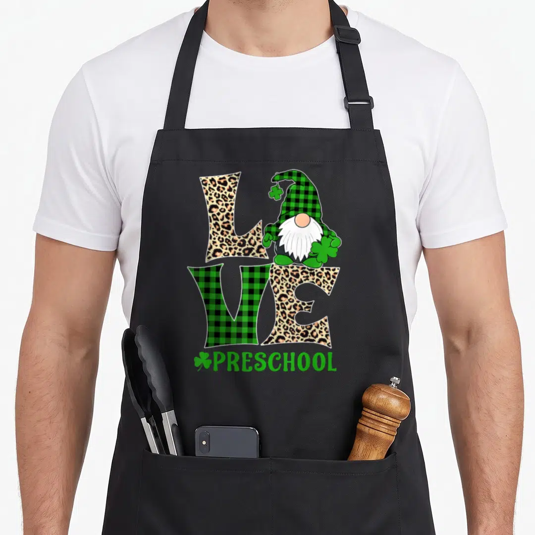 Irish preschool apron - Gnome sitting on LOVE text with PRESCHOOL and shamrock in green plaid and leopard print — shown on a black cotton canvas apron against a white background