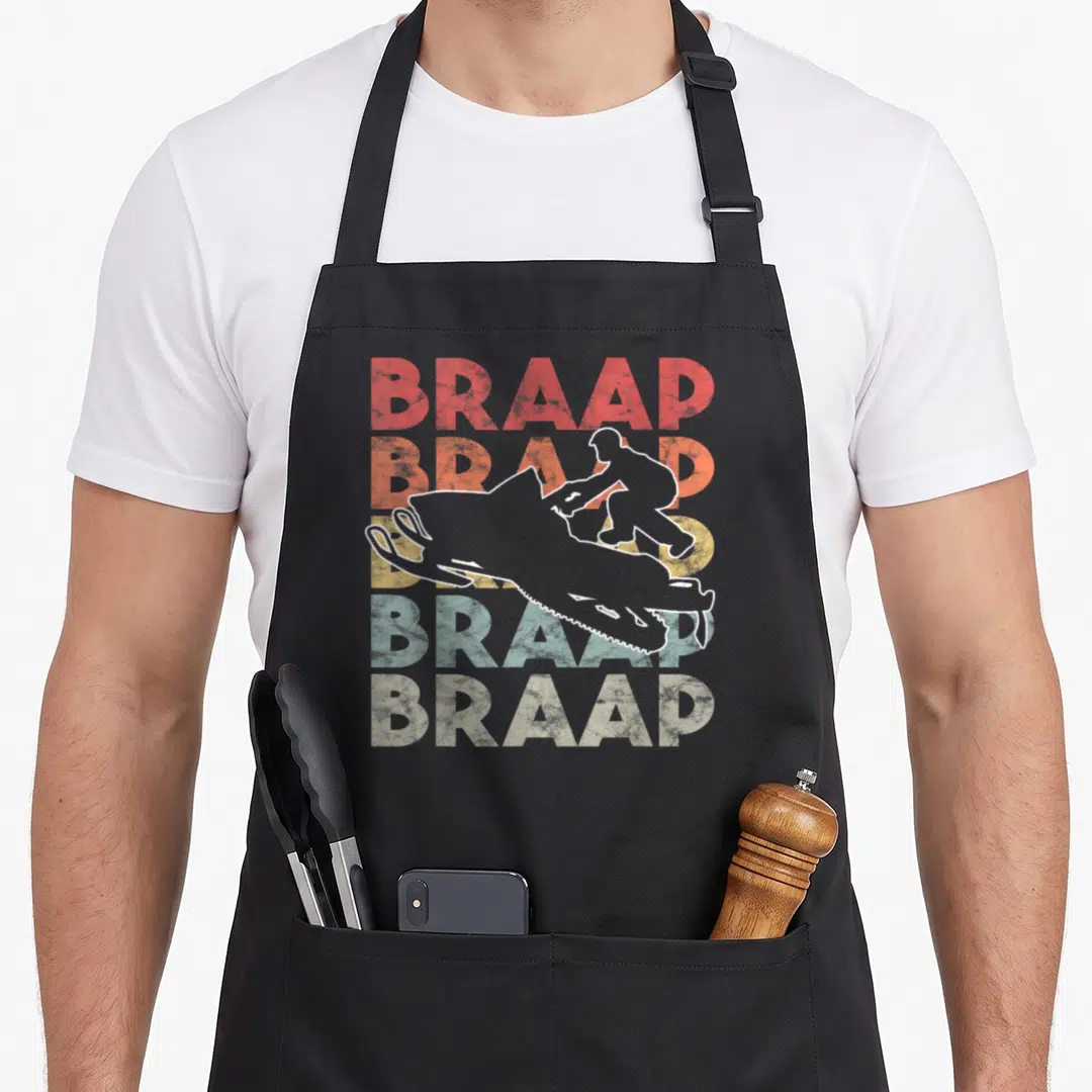 Braap snowmobile girl apron - Retro snowmobile rider silhouette over five stacked rainbow BRAAP text lines — shown on a black cotton canvas apron against a white background