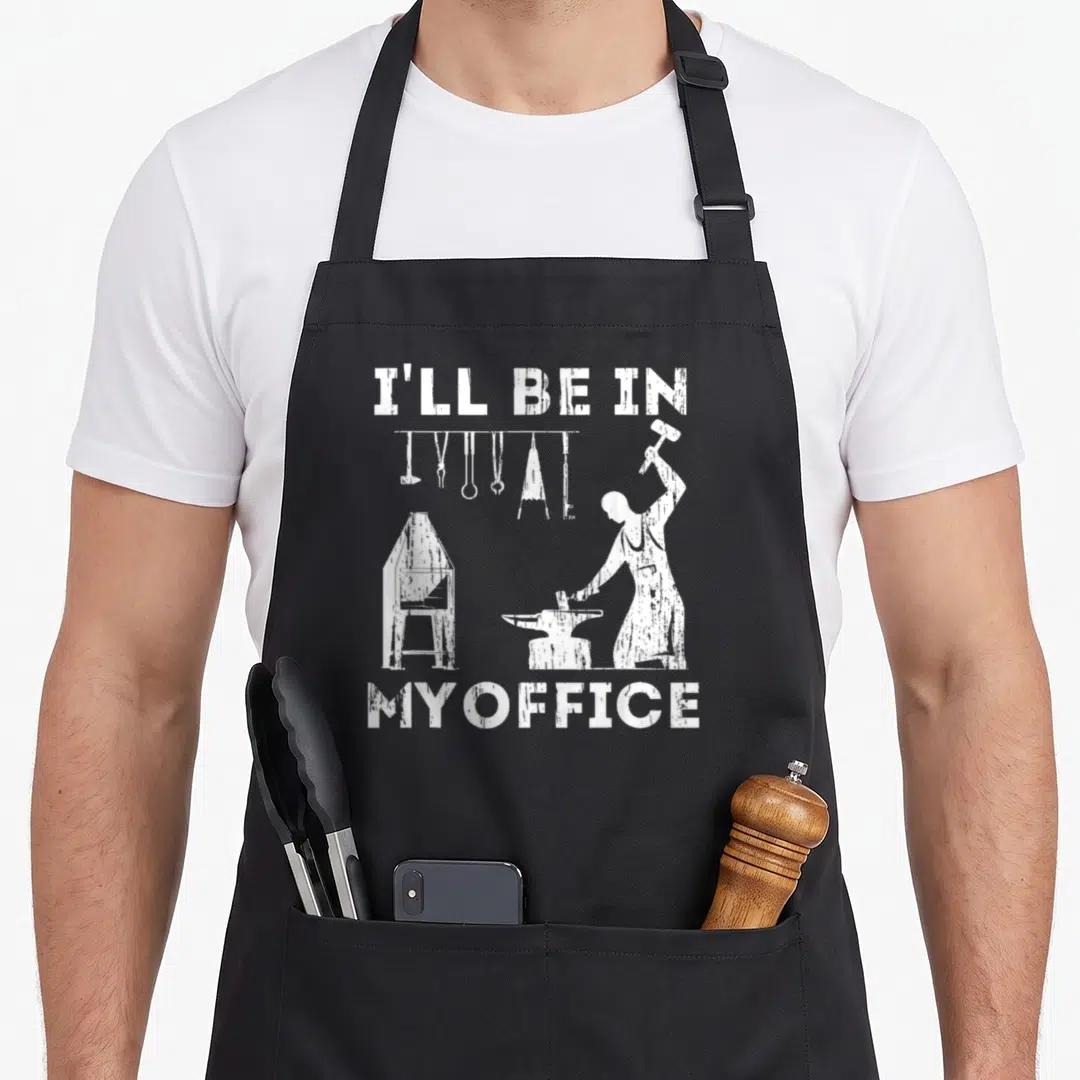 Ill blacksmith apron - White distressed text I'll Be In My Office above a blacksmith at an anvil with hanging tools — shown on a black cotton canvas apron against a white background