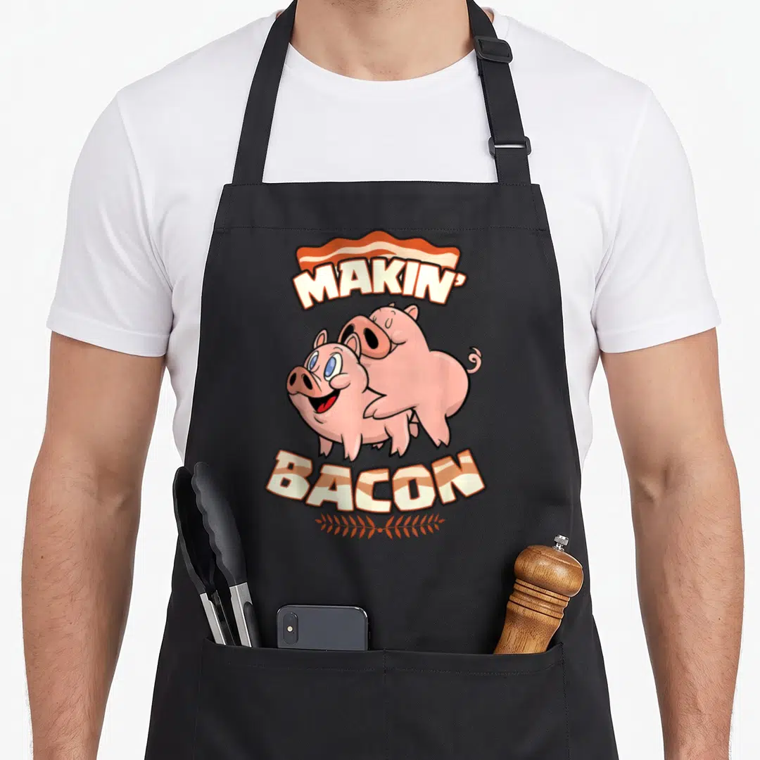 Makin bacon apron - Two pink cartoon pigs under MAKIN BACON text with bacon strip and leaf branch — shown on a black cotton canvas apron against a white background