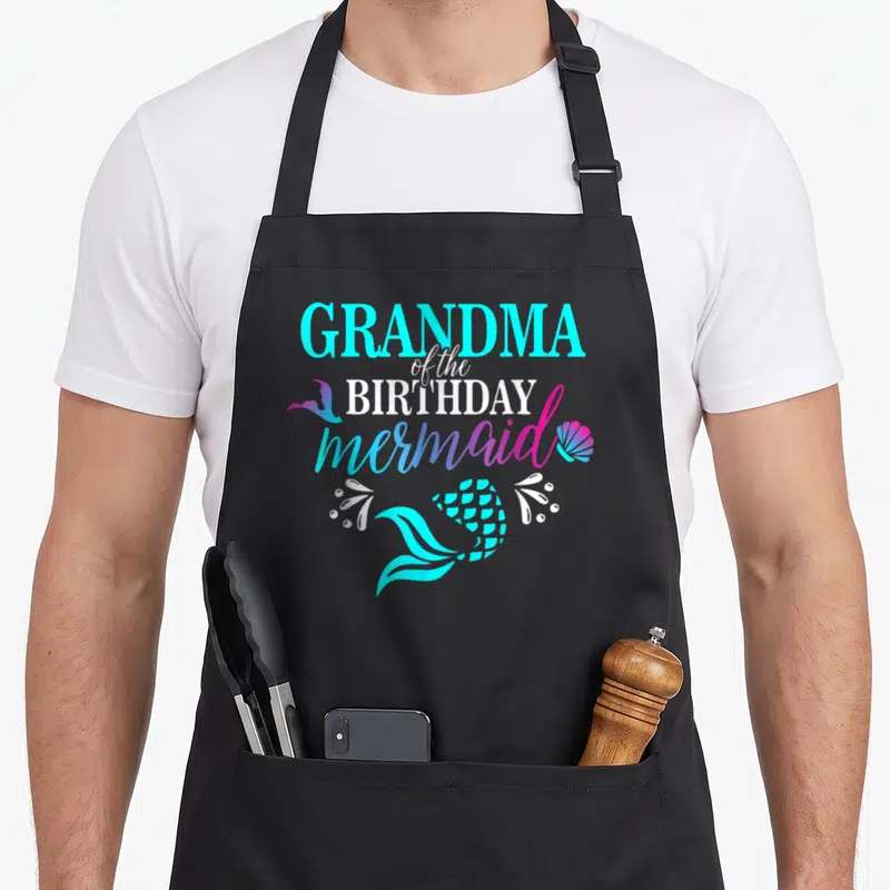 Mermaid family grandma in apron - Stacked GRANDMA of the BIRTHDAY mermaid text with cyan tail, pink seashell, and white — shown on a black cotton canvas apron against a white background