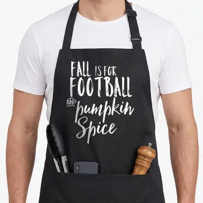 Football spice apron - White stacked typography reading FALL IS FOR FOOTBALL AND pumpkin Spice — shown on a black cotton canvas apron against a white background