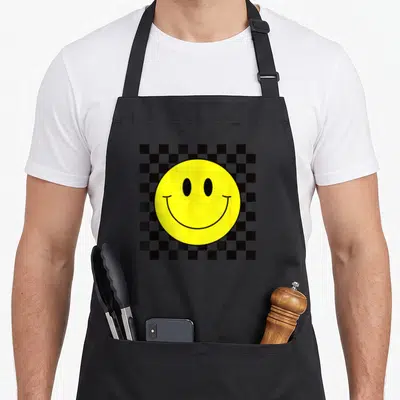 70s smile face on apron - Yellow smiley face circle over black and grey geometric checkered square pattern — shown on a black cotton canvas apron against a white background