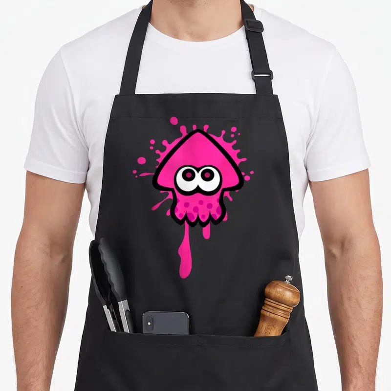 Inkling squid apron - Pink cartoon inkling head with large eyes on a pink ink splatter with a downward drip and black — shown on a black cotton canvas apron against a white background