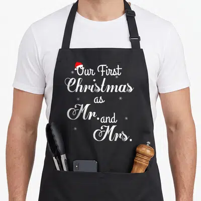 Couple wife christmas aprons - White stacked text Our First Christmas as Mr and Mrs topped with red Santa hat — shown on a black cotton canvas apron against a white background