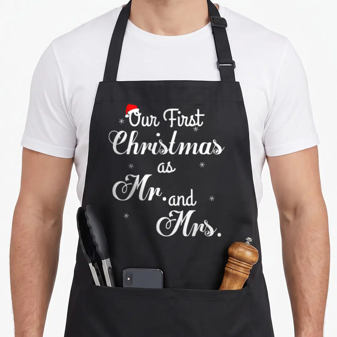 Couple wife christmas aprons - White stacked text Our First Christmas as Mr and Mrs topped with red Santa hat — shown on a black cotton canvas apron against a white background