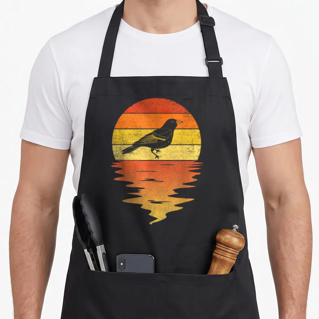 Sunset bird apron - Grey crow silhouette against red orange yellow striped sun with wavy reflection — shown on a black cotton canvas apron against a white background