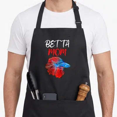 Cute betta fish apron - Stacked BETTA MOM text above a red and blue cartoon betta fish — shown on a black cotton canvas apron against a white background