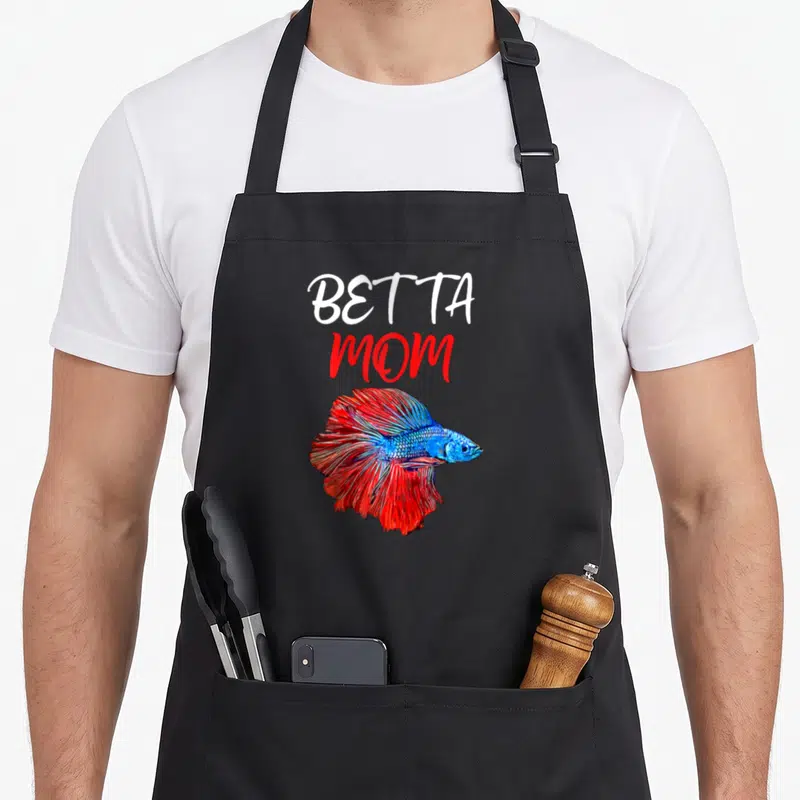 Cute betta fish apron - Stacked BETTA MOM text above a red and blue cartoon betta fish — shown on a black cotton canvas apron against a white background