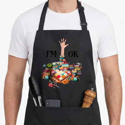 Sewing knitting apron - I'M OK text split by arm rising from colorful fabric spools and quilt cartoon style — shown on a black cotton canvas apron against a white background