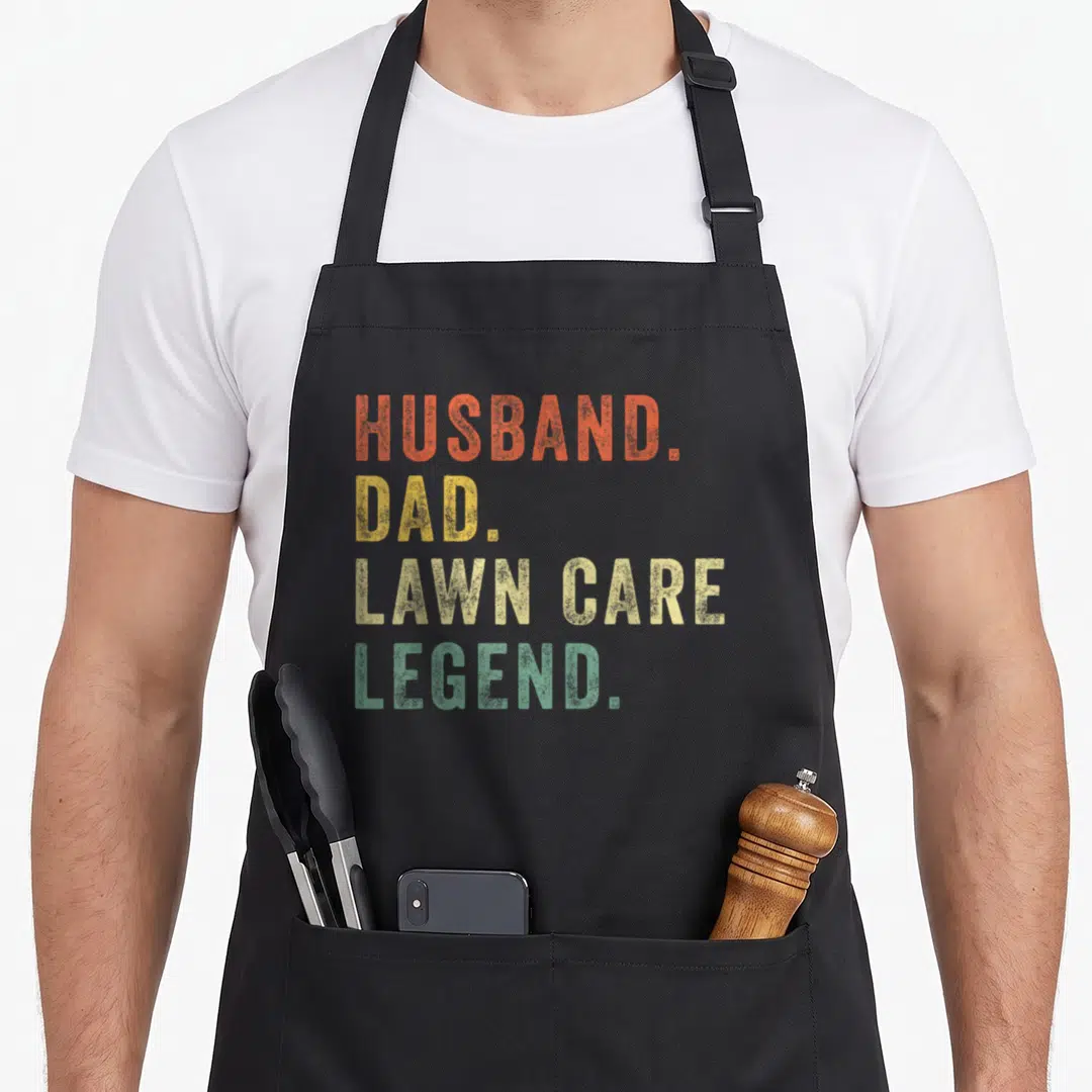 Lawn landscaper apron - Stacked HUSBAND. DAD. LAWN CARE LEGEND. text in orange, yellow, beige, and green distressed font — shown on a black cotton canvas apron against a white background