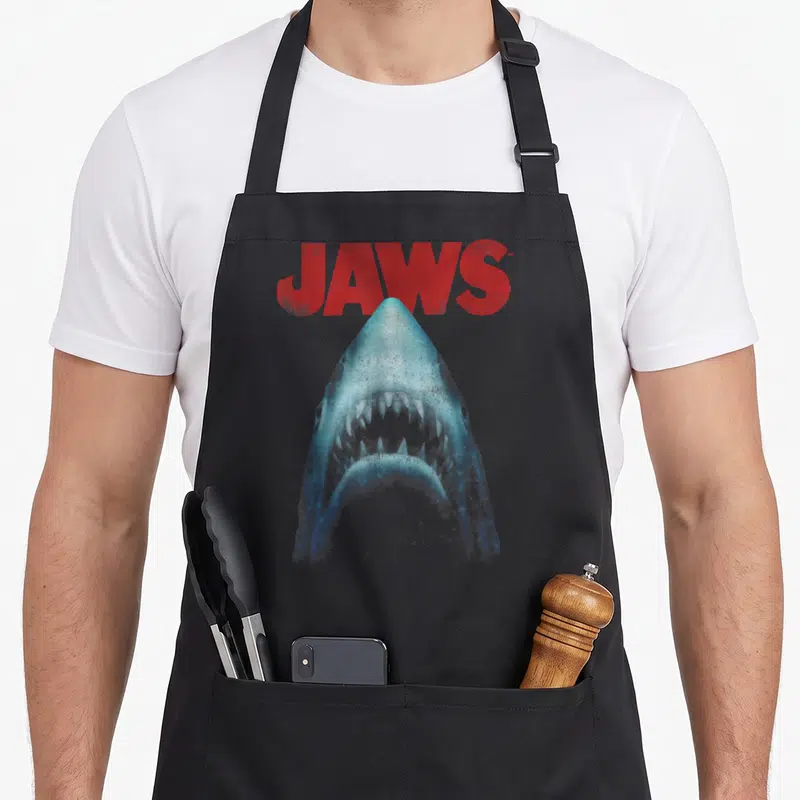 Close movie poster apron - Distressed red JAWS text above a blue shark head with open mouth pointing upward on grey — shown on a black cotton canvas apron against a white background