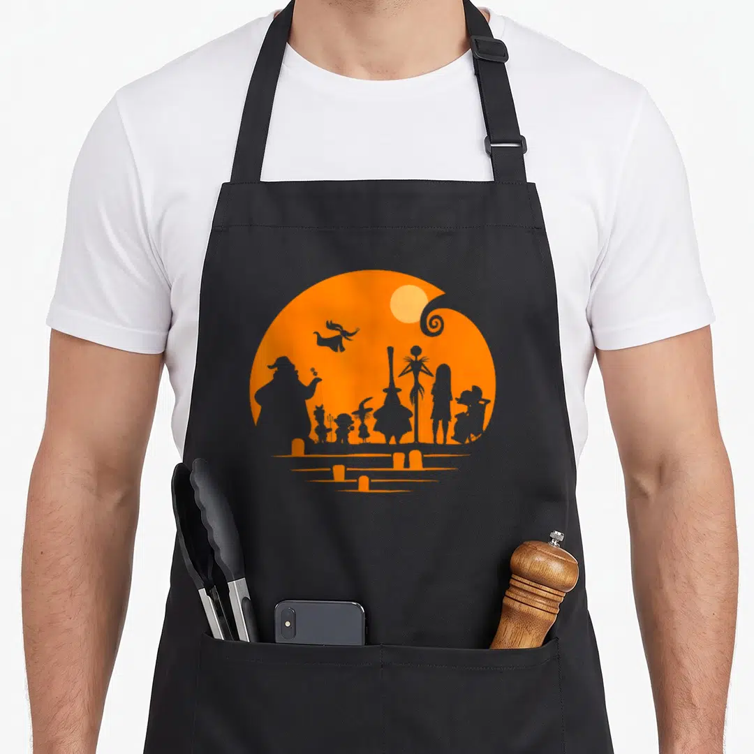 Cute silhouette halloween apron - Orange circle with character silhouettes and tombstone rows in minimalist style — shown on a black cotton canvas apron against a white background