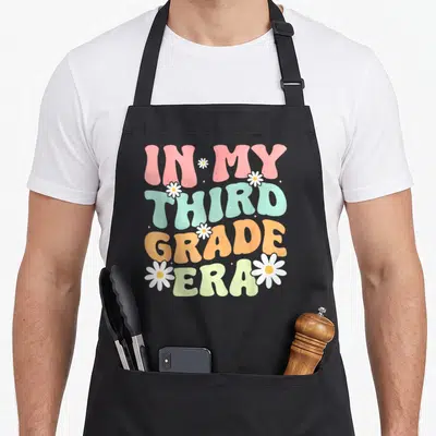 3rd groovy grade apron - Stacked IN MY THIRD GRADE ERA text in pink mint orange and pale yellow with white daisies — shown on a black cotton canvas apron against a white background