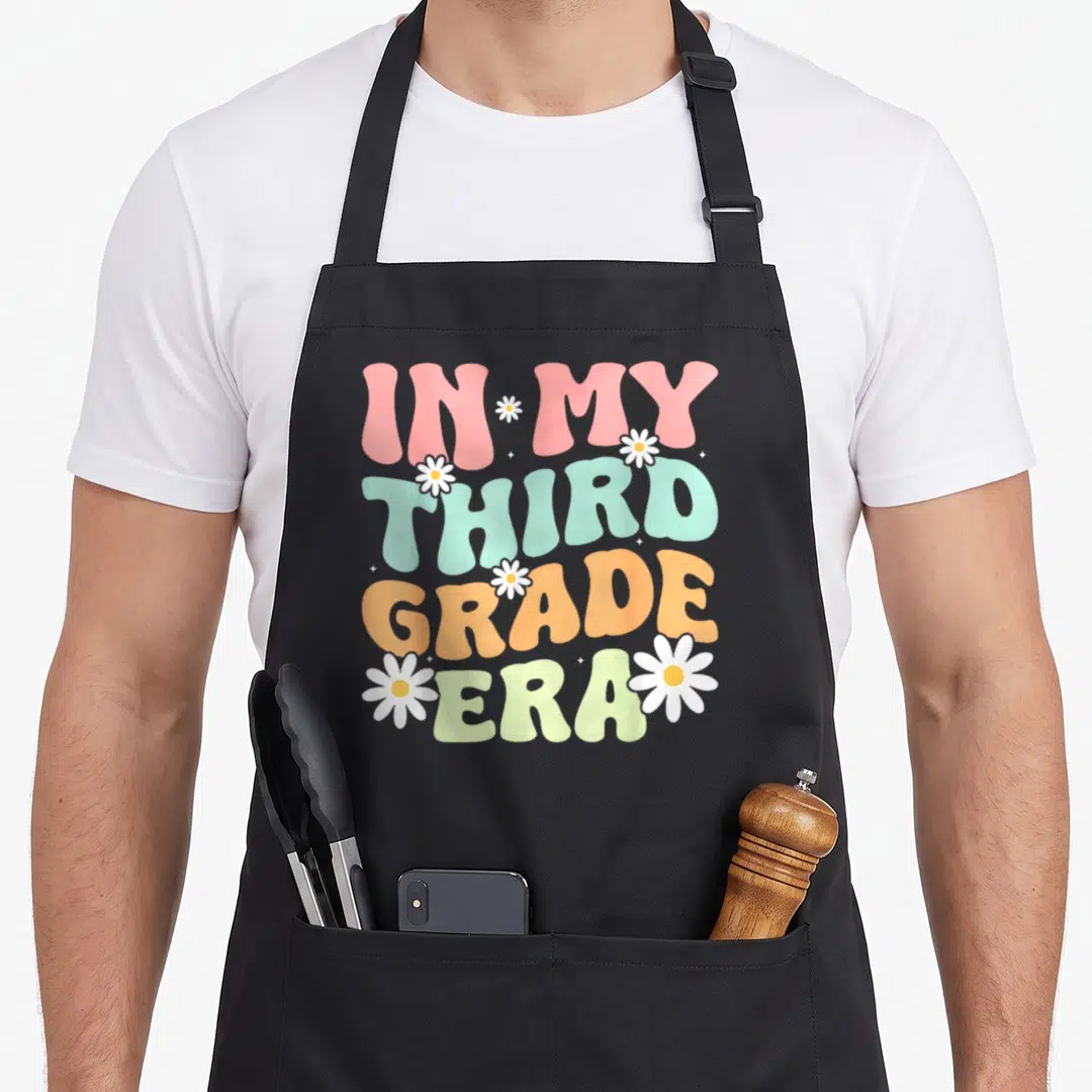 3rd groovy grade apron - Stacked IN MY THIRD GRADE ERA text in pink mint orange and pale yellow with white daisies — shown on a black cotton canvas apron against a white background