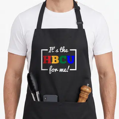 Hbcu student aprons - Stacked It's the HBCU for me text in red yellow green blue and white bracketed typography — shown on a black cotton canvas apron against a white background