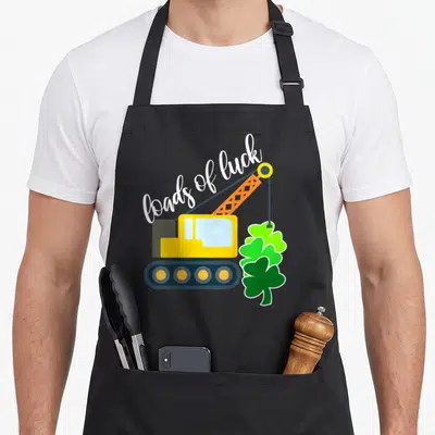 Load truck apron - Yellow crane lifting three green shamrocks under white loads of luck script text — shown on a black cotton canvas apron against a white background