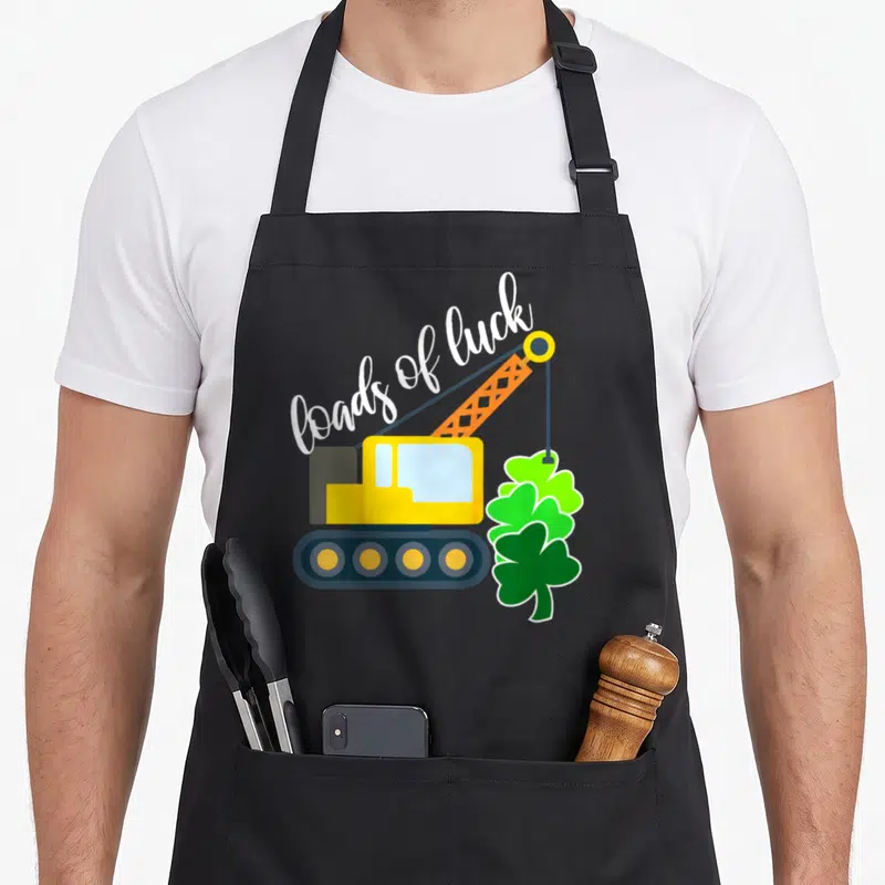Load truck apron - Yellow crane lifting three green shamrocks under white loads of luck script text — shown on a black cotton canvas apron against a white background