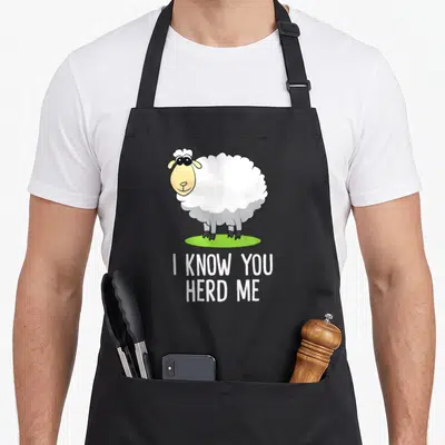 Herd sheep apron - Cartoon sheep on green grass with text I KNOW YOU HERD ME — shown on a black cotton canvas apron against a white background